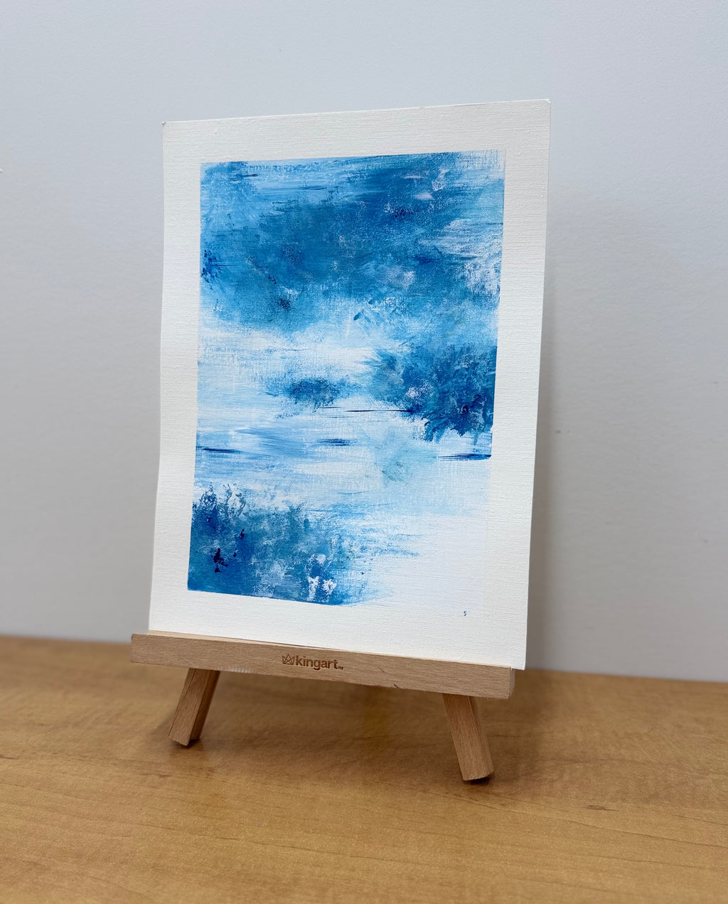 Abstract blue painting on a white canvas with a wooden easel against a light gray background.