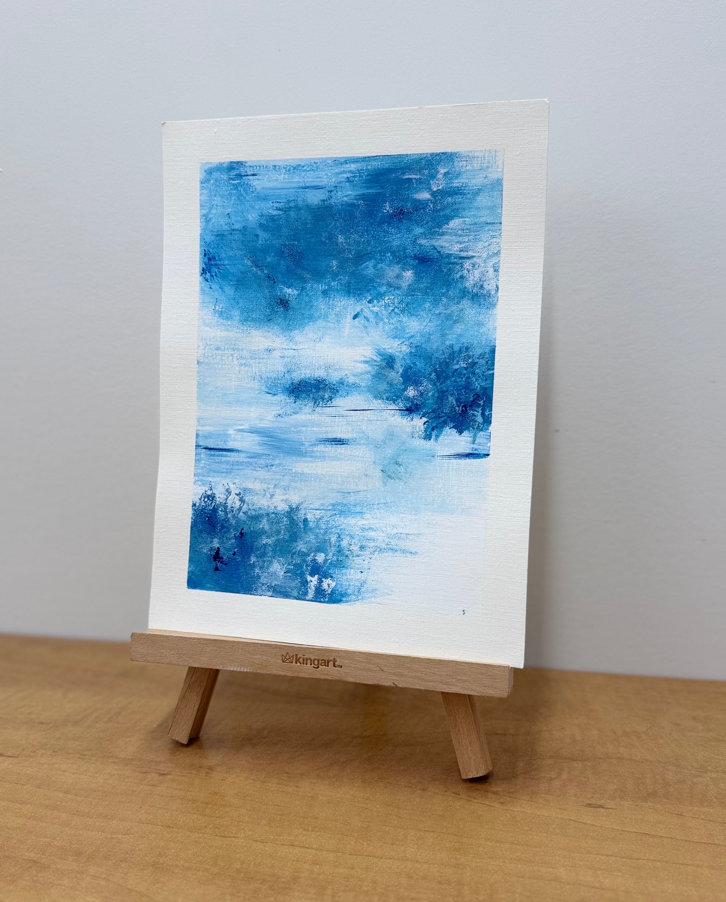 Abstract blue painting on a white canvas with a wooden easel against a light gray background.