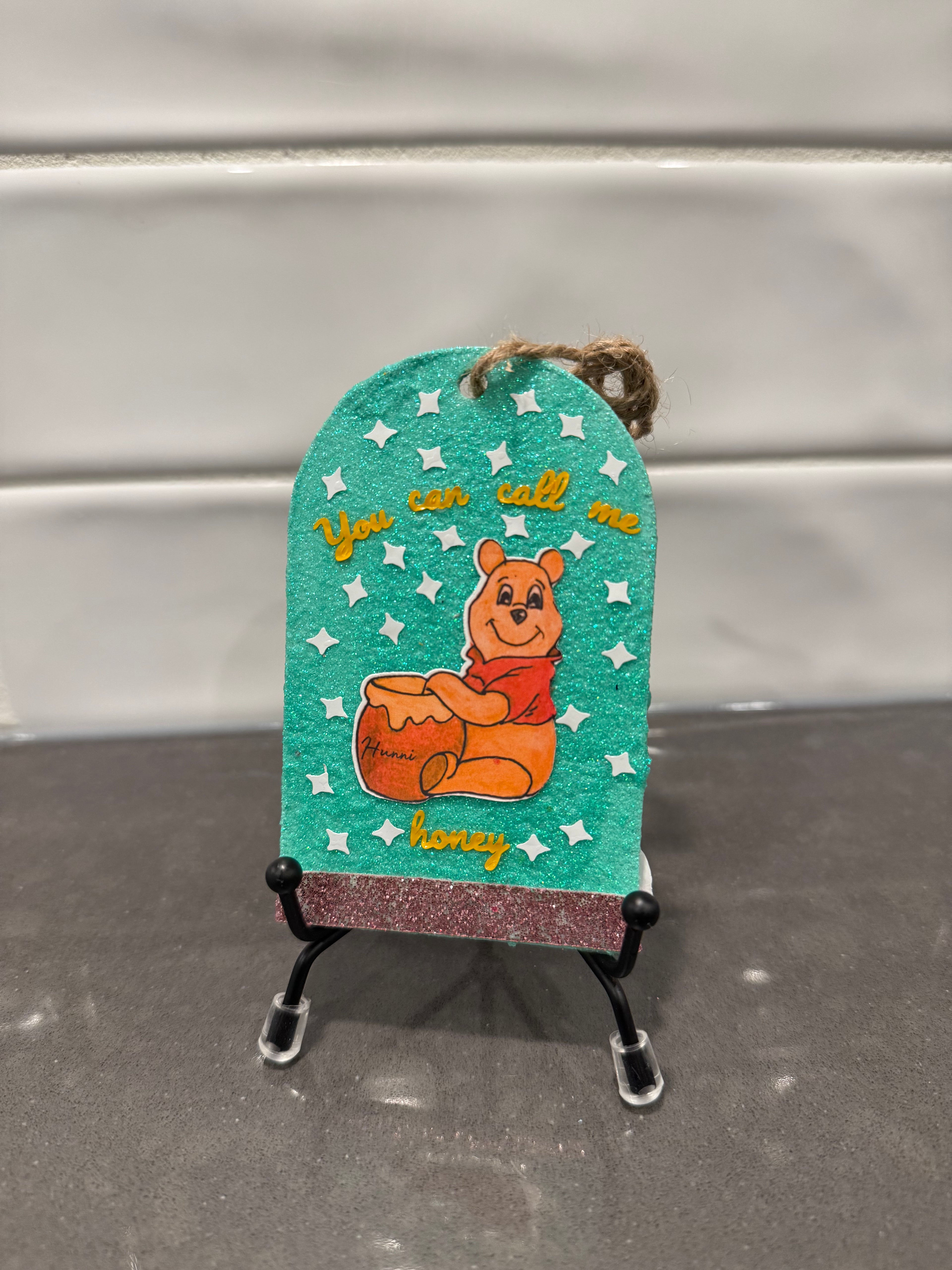 Decorative tag with a cartoon bear holding honey, on a stand against a neutral background