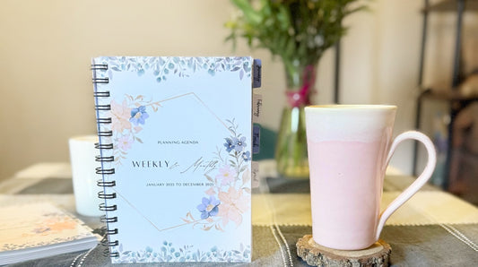 Agenda in foreground beside a pink ceramic coffee mug. Background has  flowers, book shelf against. Achieve your goals with the 2025 Empowerment Agenda. Featuring goal-setting tools, wellness trackers, inspiring quotes, and vibrant artwork.