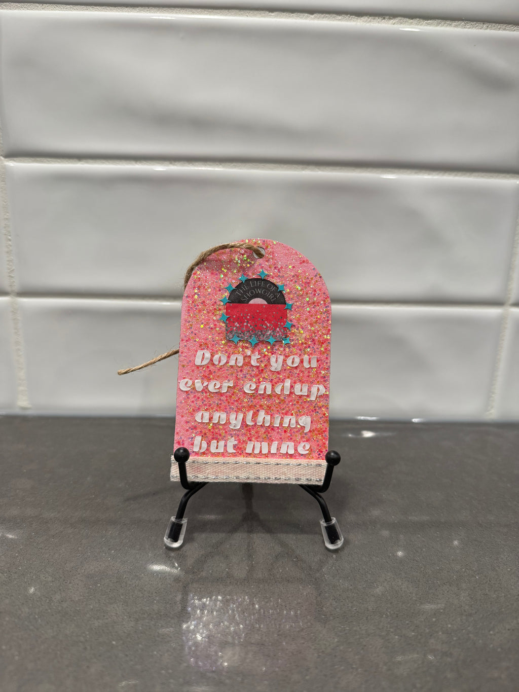 Small pink decorative item with text on a stand against a tiled wall background