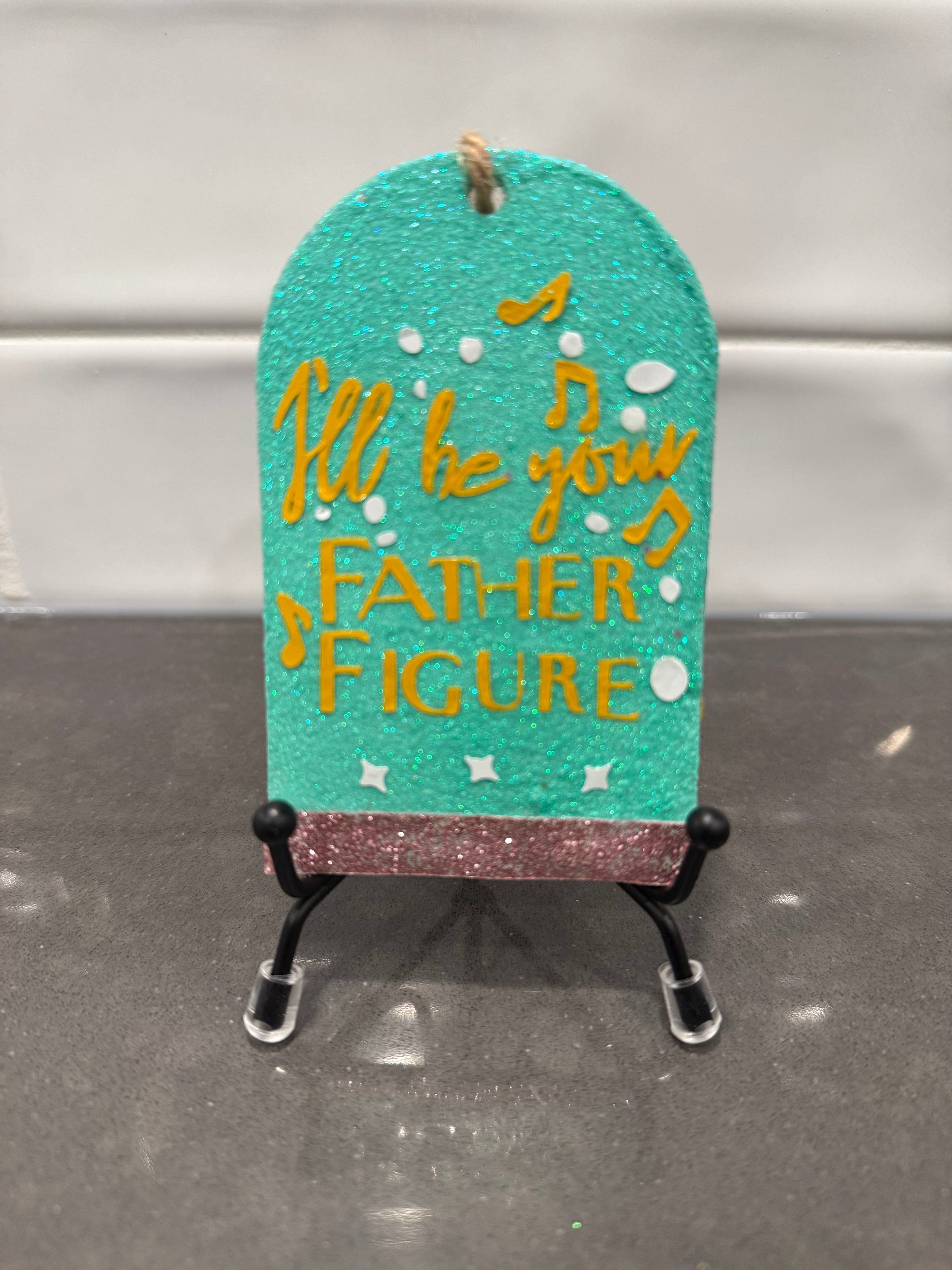 Decorative sign with 'I'll be your Father Figure' text on a stand against a neutral background