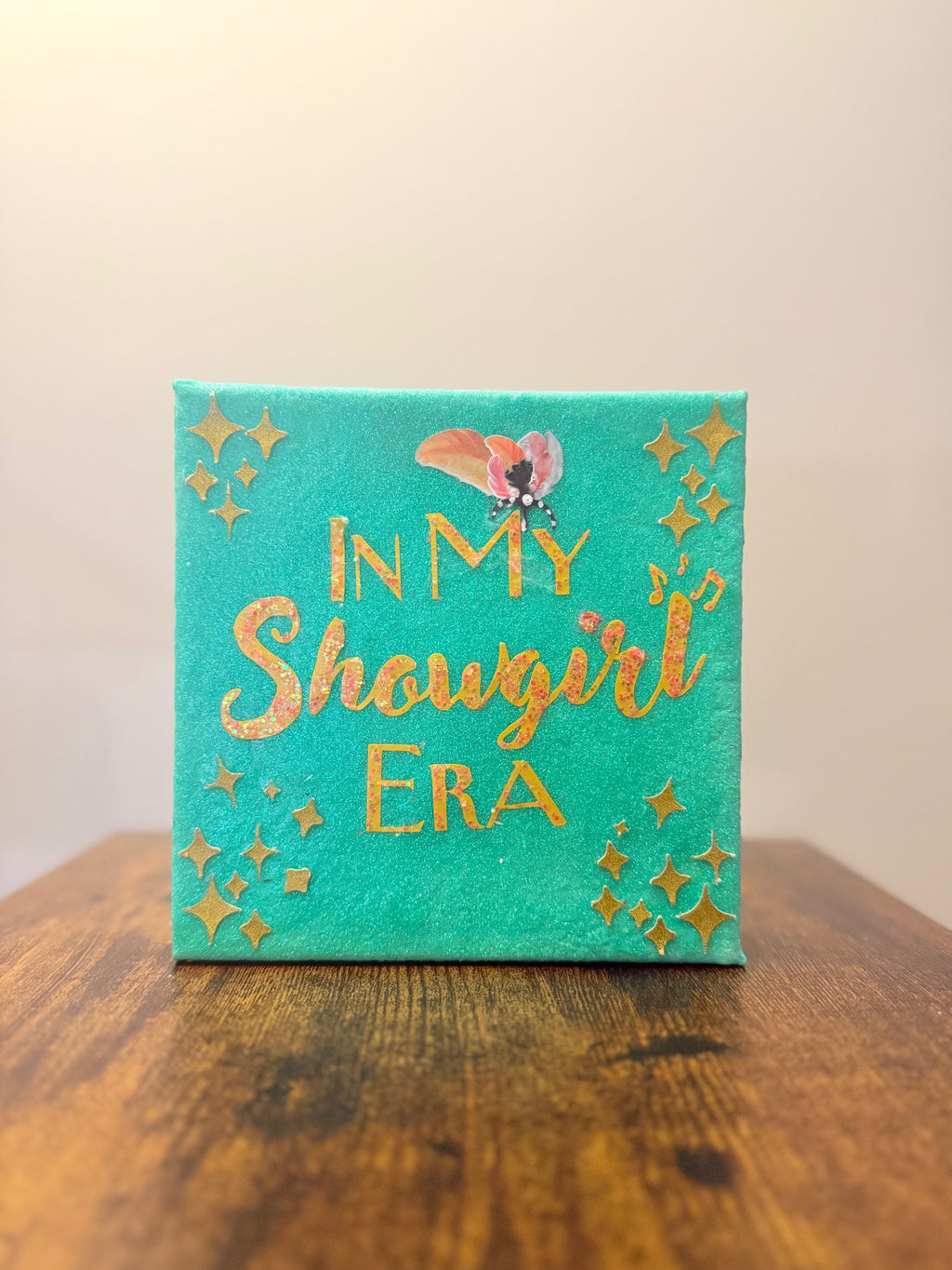 Turquoise canvas with gold text 'In My Showgirl Era' on a wooden surface.